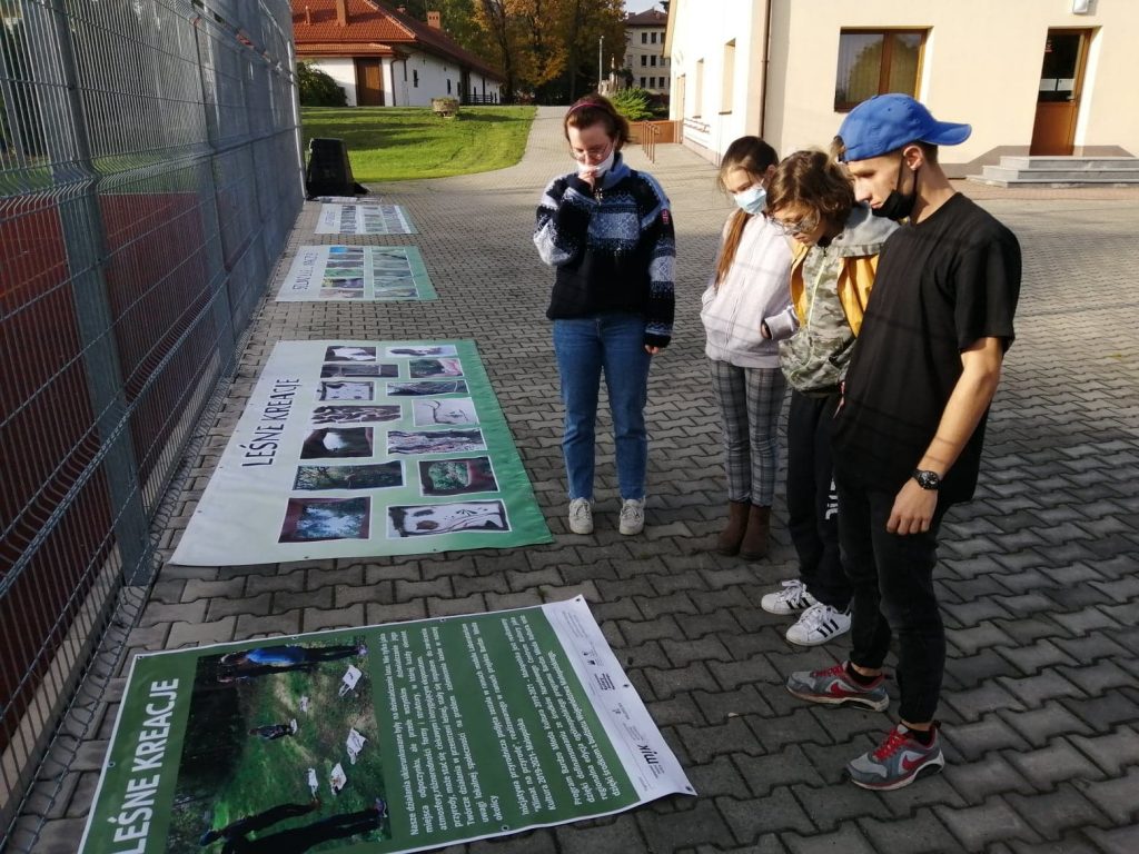 na ziemi, pod ogrodzeniem boiska sportowego leżą: plakat i trzy banery ze zdjęciami lasu, cztery osoby stoją i czytają tekst z plakatu