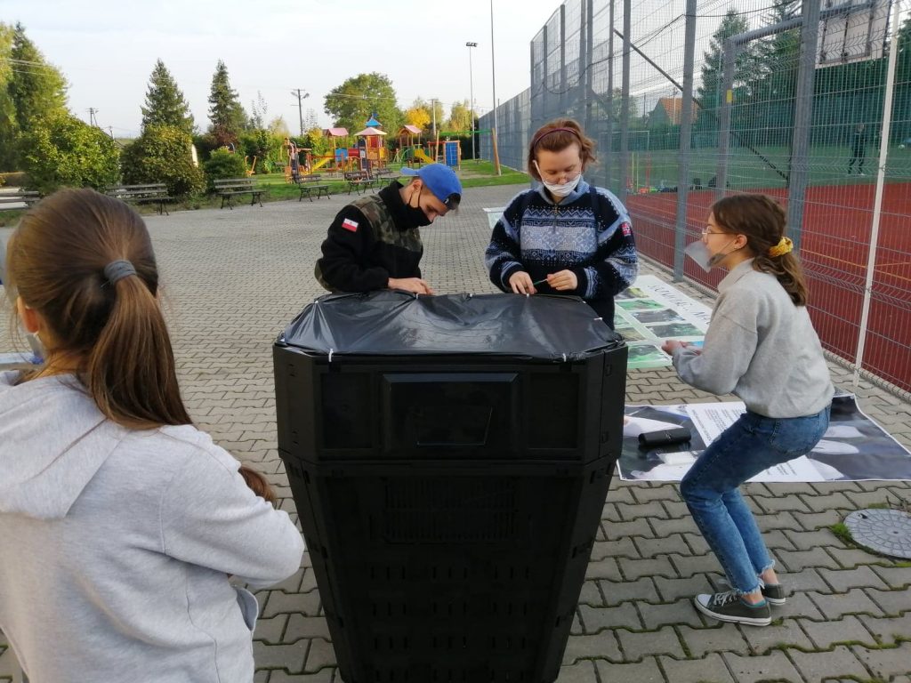 teren obok boiska sportowego, duży czarny kosz, dwie osoby przywiązują czarną folię do kosza, tak by stanowiła pokrywę kosza, obok stoją dwie inne osoby