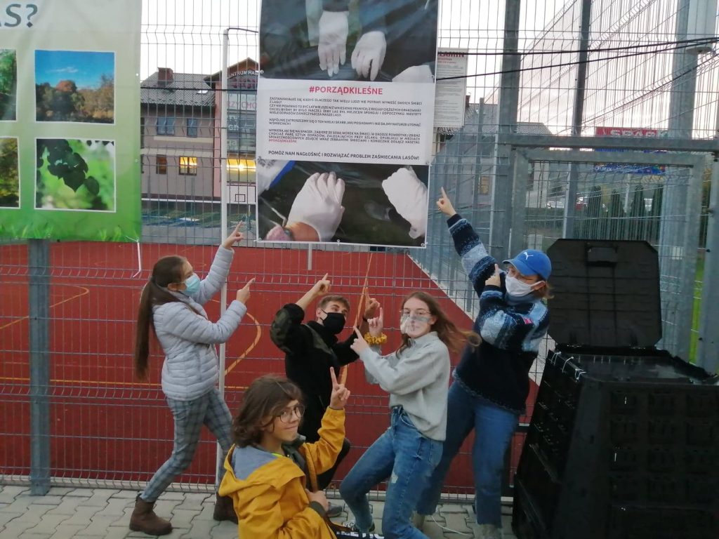 baner zawieszony na ogrodzeniu boiska sportowego, z napisem porządki leśne, pięcioro nastolatków stoi przed plakatem z uniesionymi rękami wskazującymi na plakat