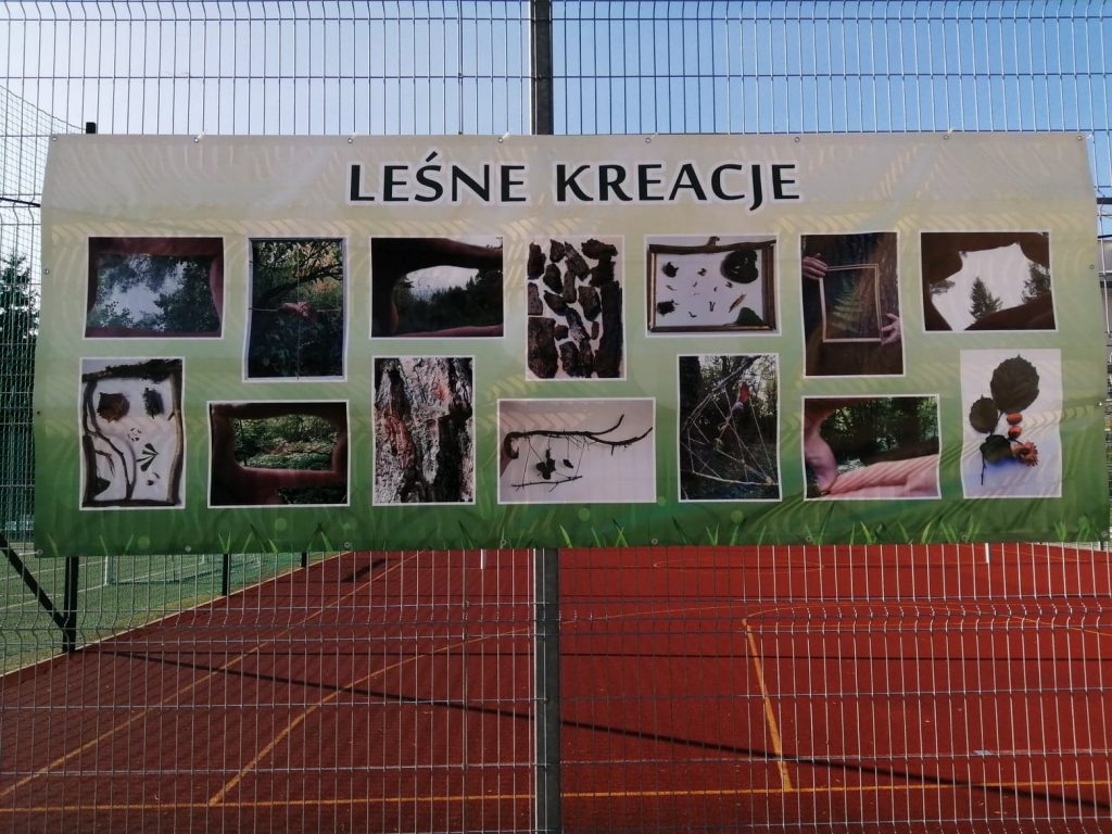 baner zawieszony na ogrodzeniu boiska sportowego z napisem Leśne Kreacje i zdjęciami przedstawiającymi w artystyczny sposób las, drzewa, gałęzie, liście, szyszki