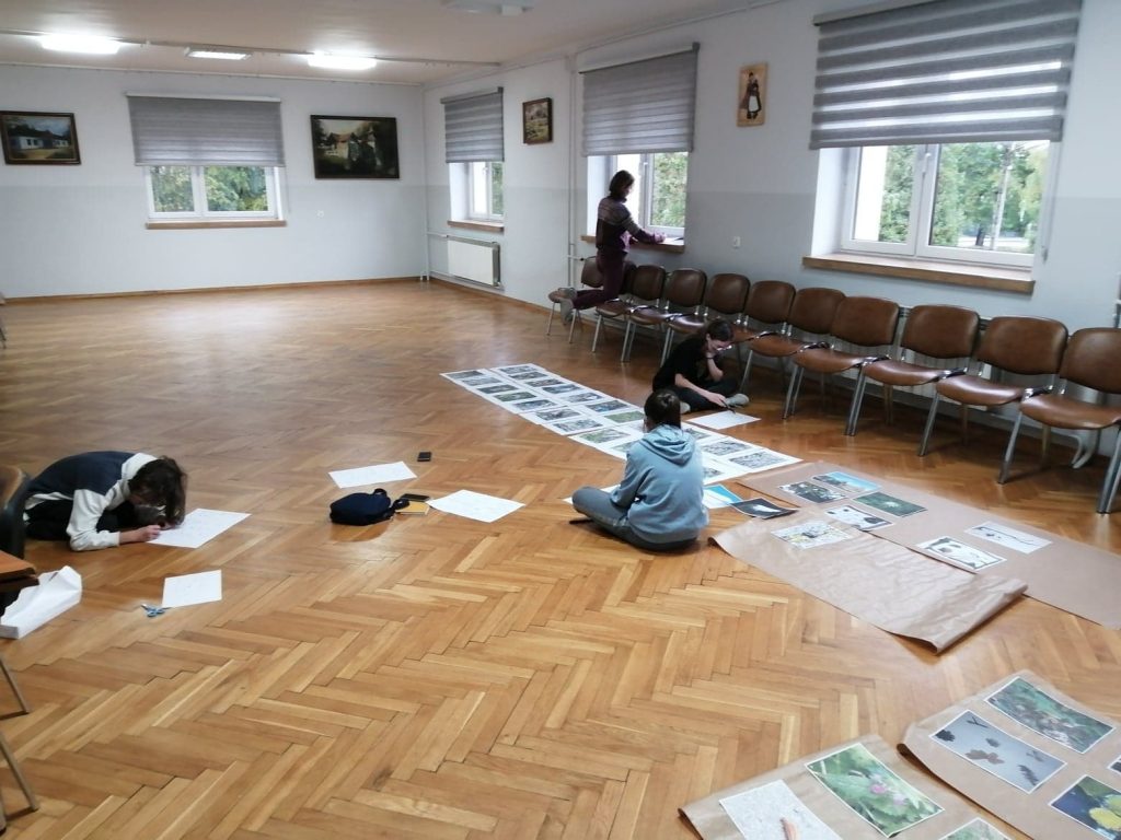 sala warsztatowa, rząd krzeseł pod oknem, na podłodze siedzi kilka dziewczynek, piszą coś na kartkach, na podłodze leżą: baner i kilka szerokich pasków szarego papieru z ułożonymi na nich zdjęciami lasu