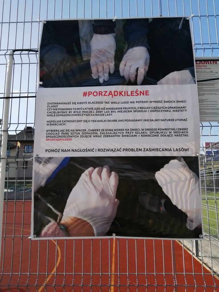 baner zawieszony na ogrodzeniu boiska sportowego, z napisem porządki leśne, tekstem opisującym problem zaśmiecania lasu, zdjęciem dłoni w rękawiczkach trzymających krawędzie czarnego kosza