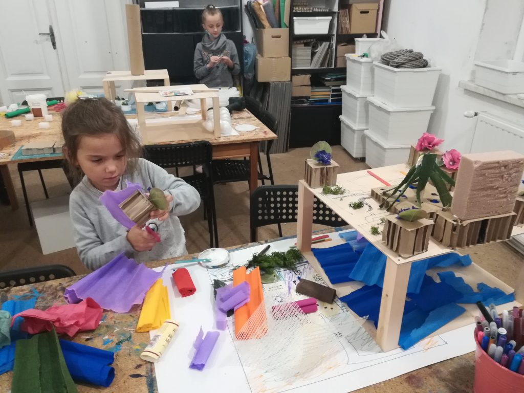 Dwie dziewczynki stoją przy stołach, na stołach modele miast stworzone z drewna, kartonu i bibuły