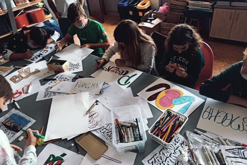 stół, na stole pisaki, kartki z graffiti, przy stole pracuje kilka młodych osób