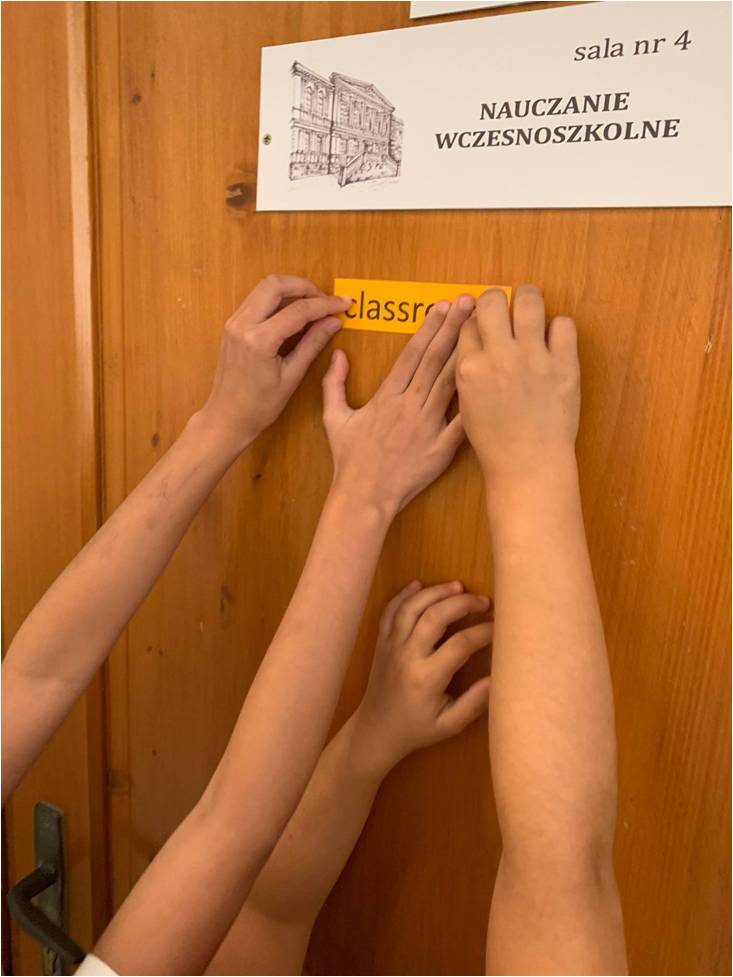 ręce przyklejają na drzwiach kartkę z napisem classroom