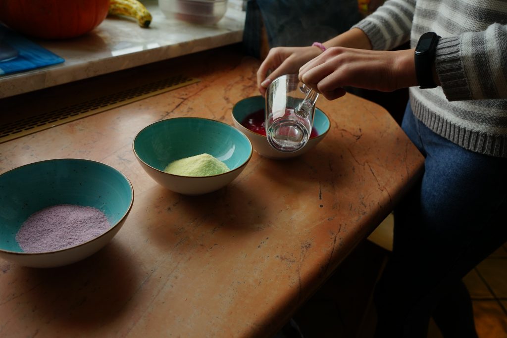 Stół, na stole zielone miski, dłonie ze szklanka z wodą wlewaną do galaretki